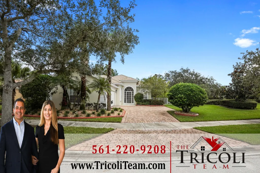 A couple in professional attire stands in a suburban driveway in front of a beige house with a large tree; promotional text for The Tricoli Team is at the bottom.