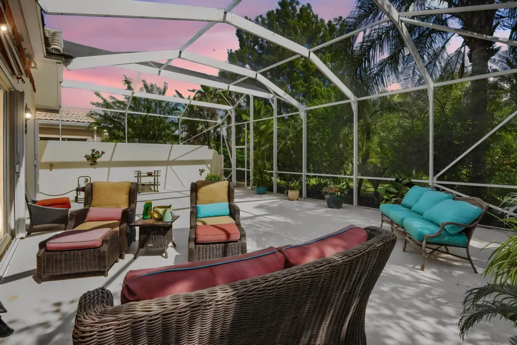 Screened outdoor patio with wicker furniture and turquoise cushions facing a white wall, pink sunset sky visible through the enclosure.