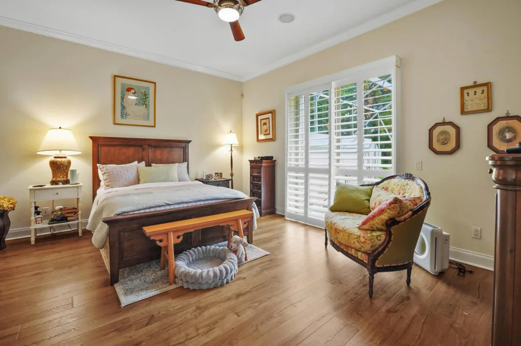 Cozy master bedroom with a wooden bed, two nightstands, lamps, and a floral upholstered chair by large white shuttered windows.