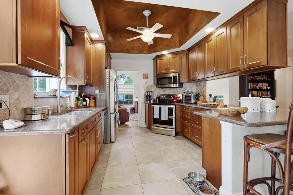 A spacious kitchen with warm wooden cabinets, granite countertops, and stainless steel appliances; a white ceiling fan hangs from a brown tray ceiling.