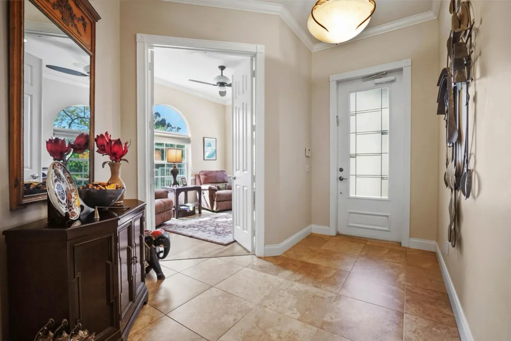 Entryway with a dark wooden console, mirror, and red flowers on a sideboard, opening into a living room with beige furniture and ceiling fan.