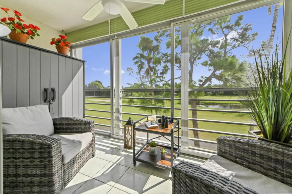 20261022153721185497000000-o - Lake Worth Homes - Lake Worth Homes Sunlit balcony with wicker loveseat and chair, a small rolling table, potted red flowers, and a view of trees and a lake beyond the railing by glass walls.