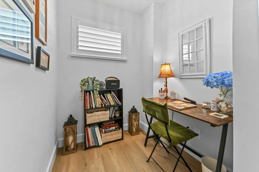 20261022153657817645000000-o - Lake Worth Homes - Lake Worth Homes Cozy home office nook with a wooden desk, green folding chair, blue hydrangeas in a vase, lamp, and a bookshelf full of books nearby.