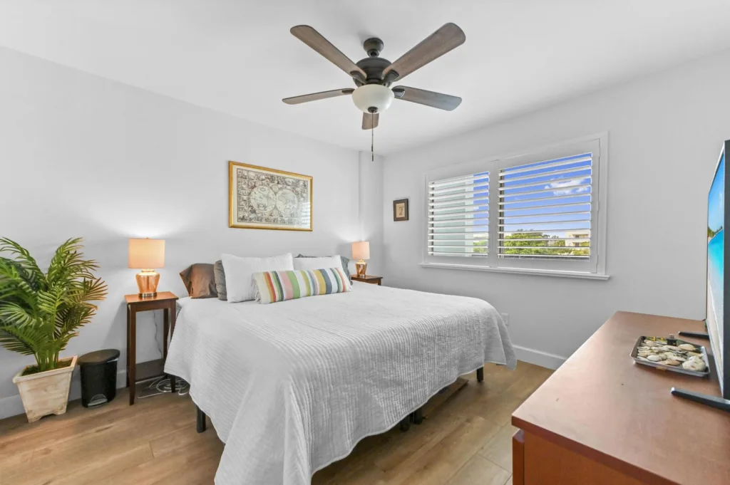 20261022153542629767000000-o - Lake Worth Homes - Lake Worth Homes Bright bedroom with a white bed, colorful striped pillow, two wooden nightstands, and warm lamps on each side.