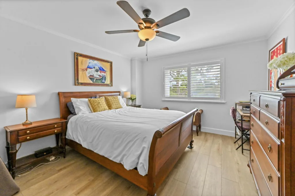 20261022153841759370000000-o - Lake Worth Homes - Lake Worth Homes Bedroom with a wooden bed flanked by nightstands, two lamps, and a dresser; window with blinds on the right side letting in light.