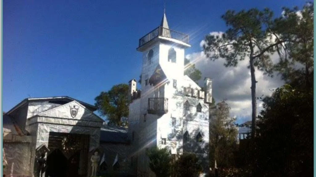 White castle-style building with a tall tower, balconies, and a gated entrance, set among trees under a blue sky.