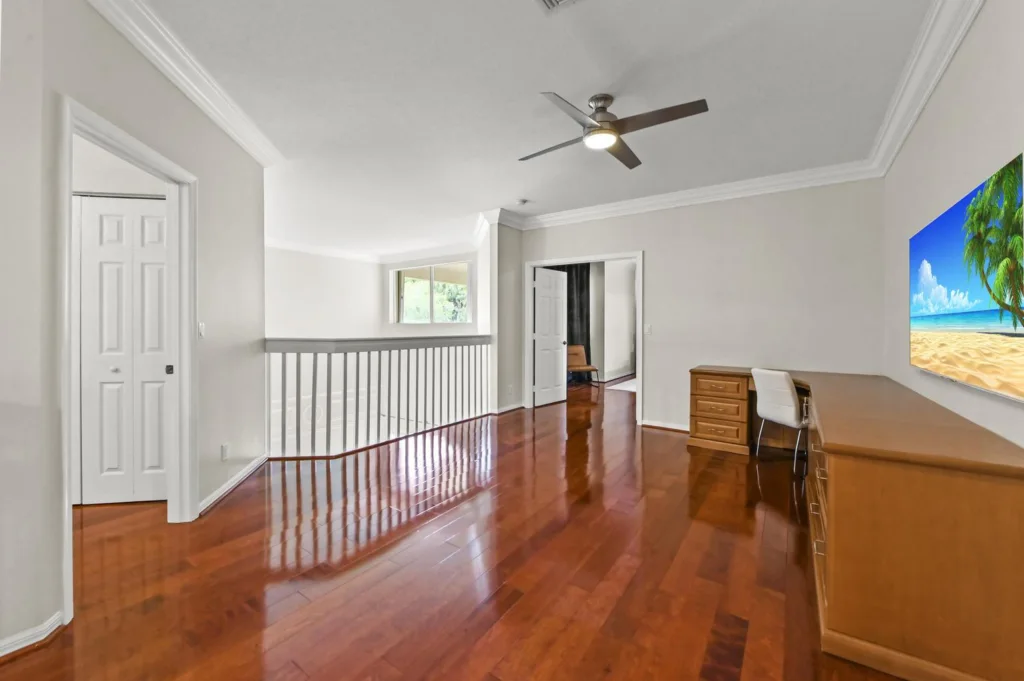 20261020152813190915000000-o - Lake Worth Homes - Lake Worth Homes Bright upstairs hallway with rich wood floors, white walls, and a ceiling fan; desk and chair against the wall, large beach image on the right.