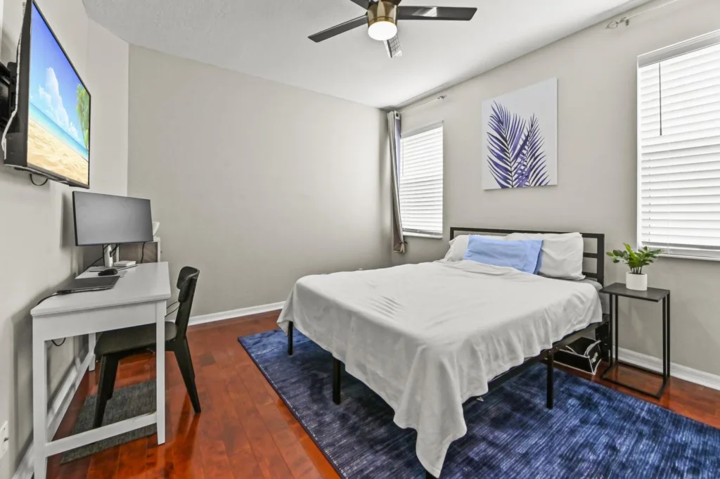 20261019172136313742000000-o - Lake Worth Homes - Lake Worth Homes Cozy bedroom with a bed, blue rug, and a white desk with a monitor setup nearby against light gray walls.