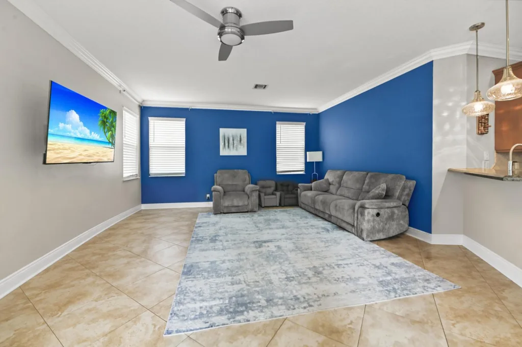 20261020152941590987000000-o 1 - Lake Worth Homes - Lake Worth Homes Living room with a blue feature wall, gray sectional and chairs, and a wall-mounted TV showing a beach scene