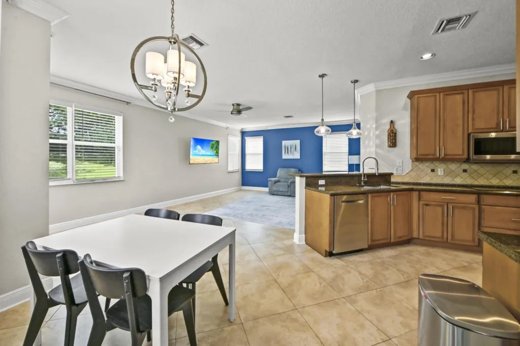 20261020152854986549000000-o - Lake Worth Homes - Lake Worth Homes Open-concept kitchen and living area with a white dining table and black chairs, a blue accent wall, and a sofa in front of a wall-mounted TV.