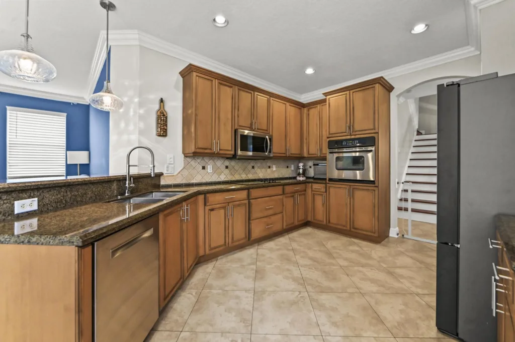 20261019171951739735000000-o - Lake Worth Homes - Lake Worth Homes U-shaped kitchen with wooden cabinets, granite countertops, and stainless steel appliances, blue accent wall on left, tiled floor.