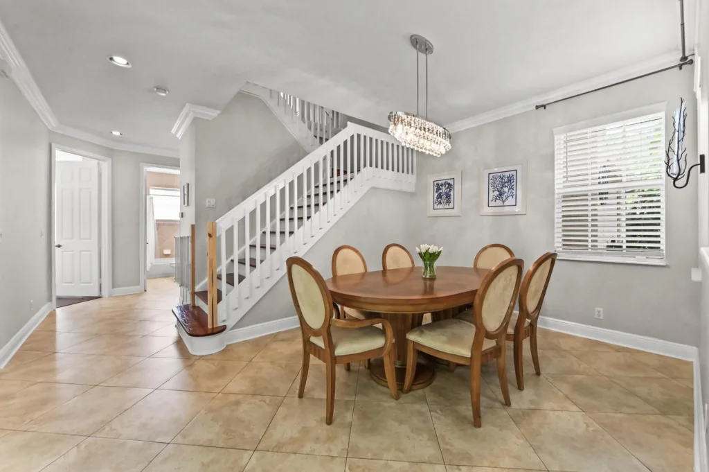20261019171905783597000000-o 1 - Lake Worth Homes - Lake Worth Homes Dining room with round wooden table and six upholstered chairs beneath a chandelier; staircase along the wall, light gray walls, and tiled floor with blinds on the window.