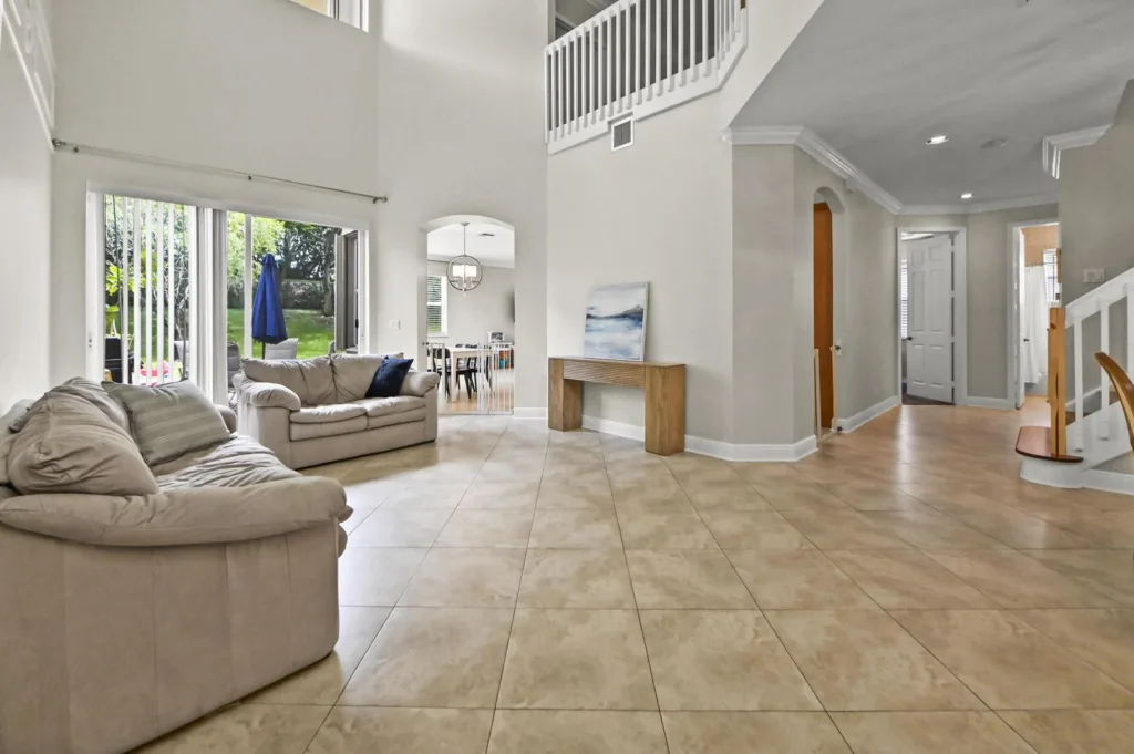 20261019171757764025000000-o - Lake Worth Homes - Lake Worth Homes Spacious living room with beige leather sofas, tiled floor, and large sliding doors opening to a green backyard with a blue patio umbrella. A wooden console with a painting sits against a wall, and a curved staircase leads upstairs nearby.